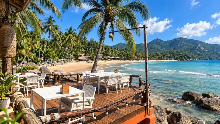 Terrace of Rim Lae Restaurant above the rocks, with white tables, palms and the bay, Koh Tao